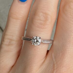Close-up of a hand wearing a solitaire diamond engagement ring on the middle finger, with a split shank band and blue nail polish on the index finger nearby.