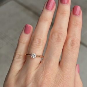 Close-up of a hand with pink nails wearing a solitaire diamond ring on a rose gold band.