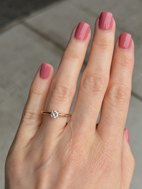 Close-up of a hand with pink nails wearing a solitaire diamond ring on a rose gold band.