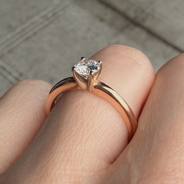 Close-up of a gold/rose-gold solitaire engagement ring on a finger, with the diamond held by prongs.