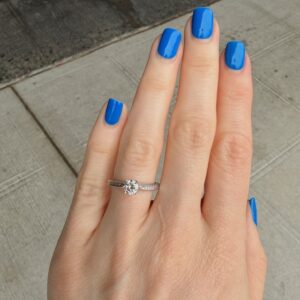 Hand with bright blue manicure shows a diamond solitaire engagement ring on the ring finger.