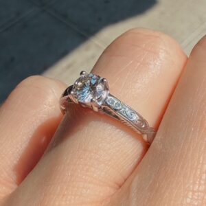 Close-up of a silver engagement ring with a large round diamond and an intricately engraved band on a finger, outdoors.