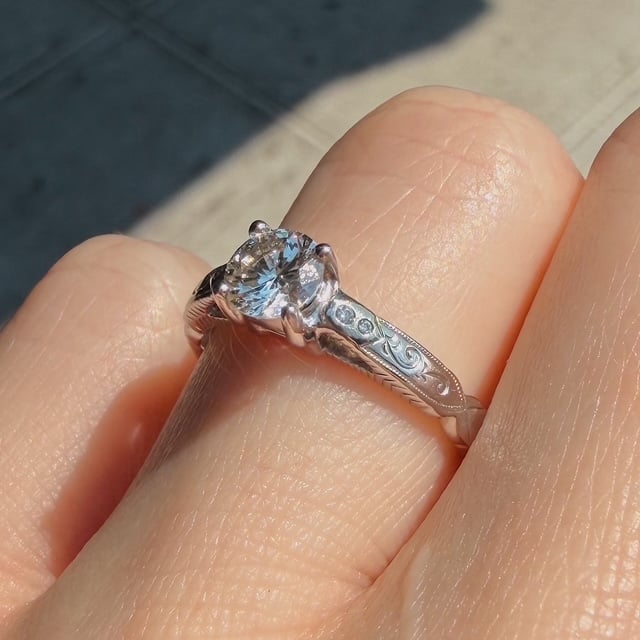 Close-up of a silver engagement ring with a large round diamond and an intricately engraved band on a finger, outdoors.