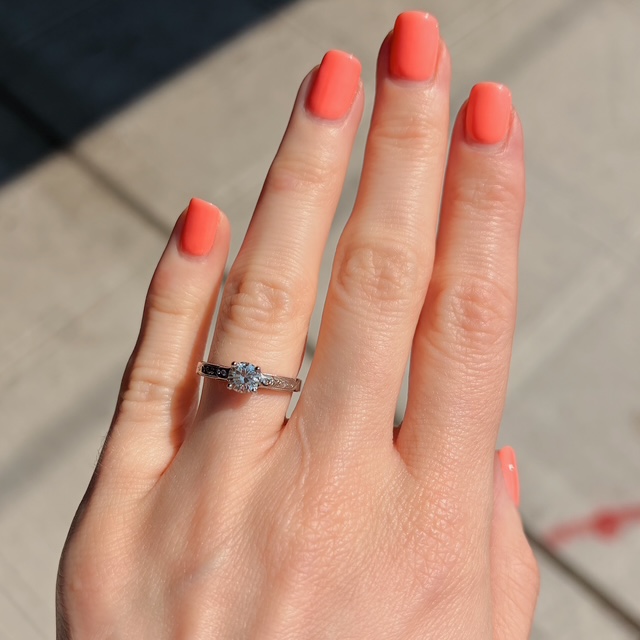 Close-up of a hand with coral nail polish wearing a diamond engagement ring on the ring finger.
