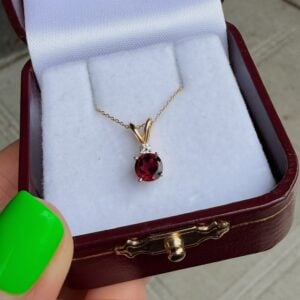 Gold pendant necklace with a red ruby and a small diamond accent, displayed in a burgundy jewelry box.