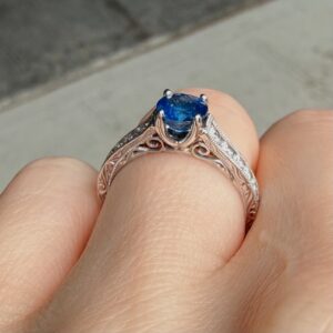 Rose-gold ring with an ornate filigree band and a deep blue oval gemstone held between fingers, close-up view