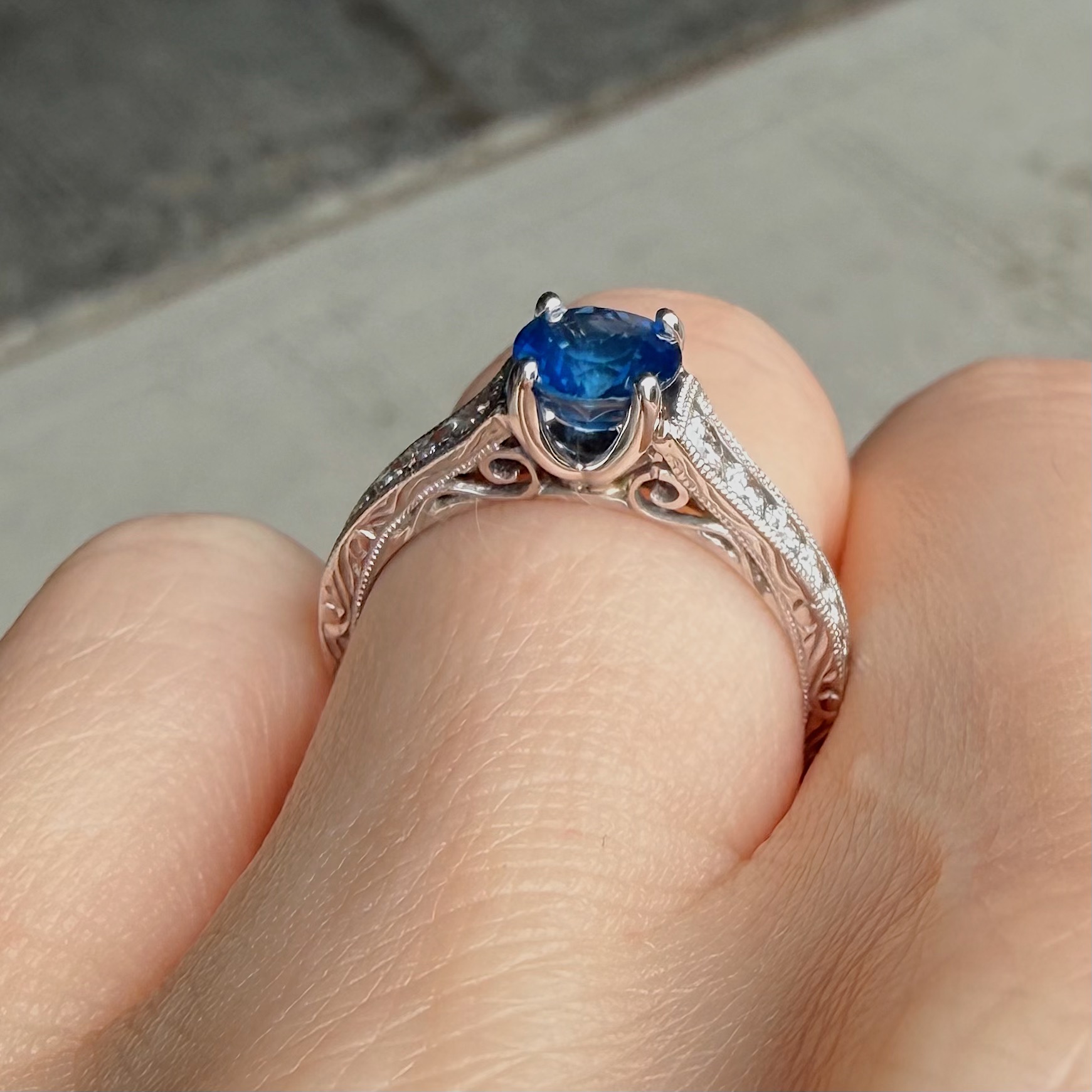 Rose-gold ring with an ornate filigree band and a deep blue oval gemstone held between fingers, close-up view