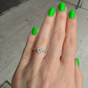 Hand with neon green nail polish wearing a diamond engagement ring on the ring finger, outdoors on a sidewalk.