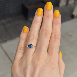 Close-up of a hand with bright yellow nails wearing a gold ring featuring a blue sapphire center stone flanked by two diamonds on each side.