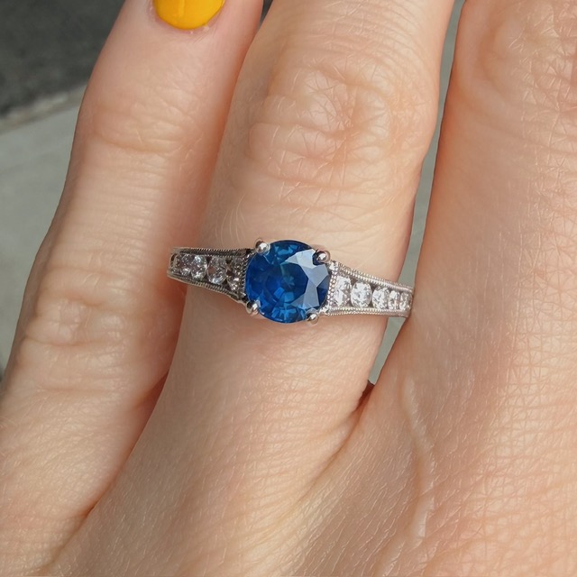 Silver ring with a blue oval-cut center stone and pavé diamonds along the band on a finger with yellow nail polish.
