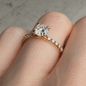 Close-up of a rose-gold engagement ring with a round center diamond and a pavé diamond band worn on a finger.