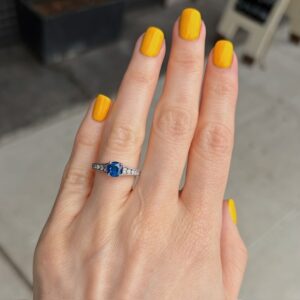 Hand with bright yellow nail polish wearing a silver ring with a blue oval gemstone and small diamonds on the band.