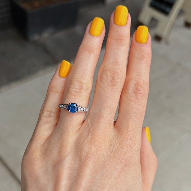 Hand with bright yellow nail polish wearing a silver ring with a blue oval gemstone and small diamonds on the band.