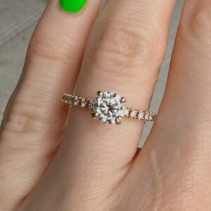 Diamond engagement ring with a round center diamond on a gold band set with small diamonds along the band, worn on a finger with bright green nail polish.