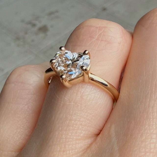 Gold solitaire diamond engagement ring on a finger with a two-tone skin tone background.