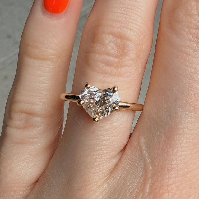 Gold engagement ring with a heart-shaped diamond on the ring finger, worn on a hand with orange nail polish.