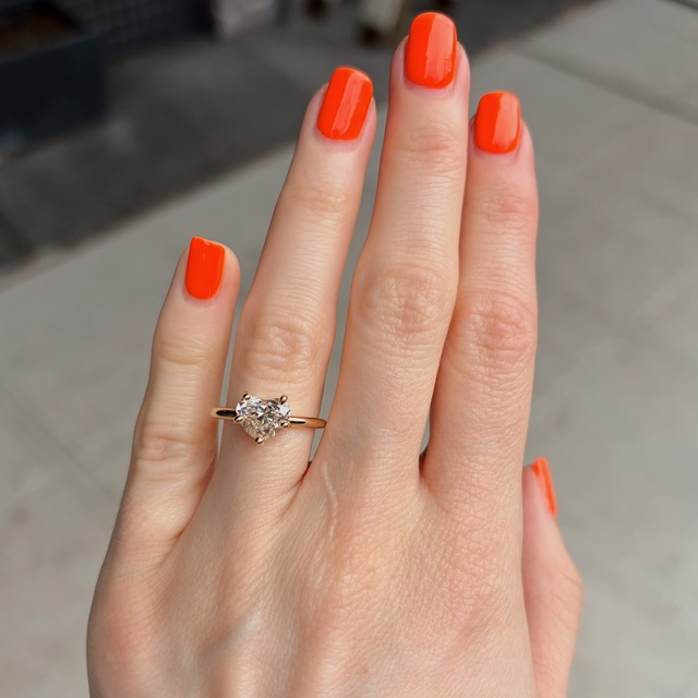 Close-up of a hand with bright orange manicure wearing a gold engagement ring with a diamond solitaire.