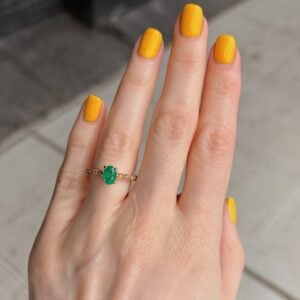 Hand with bright yellow nails wearing a gold ring with a green emerald centerpiece and small diamonds on the band, shown on the ring finger.