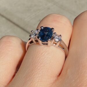 Ring with a blue oval center gemstone flanked by two small diamonds on a rose-gold band worn on a finger