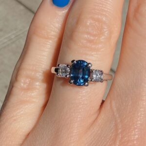 Engagement ring with a blue oval gemstone flanked by two diamonds on a white gold band on a finger.
