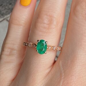 Close-up of a finger wearing a rose-gold ring with a large oval emerald-cut green gem held by four prongs, and a diamond-accent band.