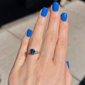 Hand with bright blue manicure wearing a blue oval gemstone ring flanked by small diamonds on a silver band (close-up)