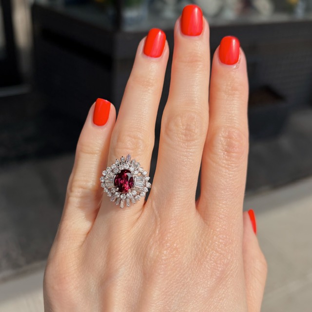 Hand with bright red manicure wearing a diamond-ring featuring a central red gemstone with a halo of smaller diamonds on the ring finger.