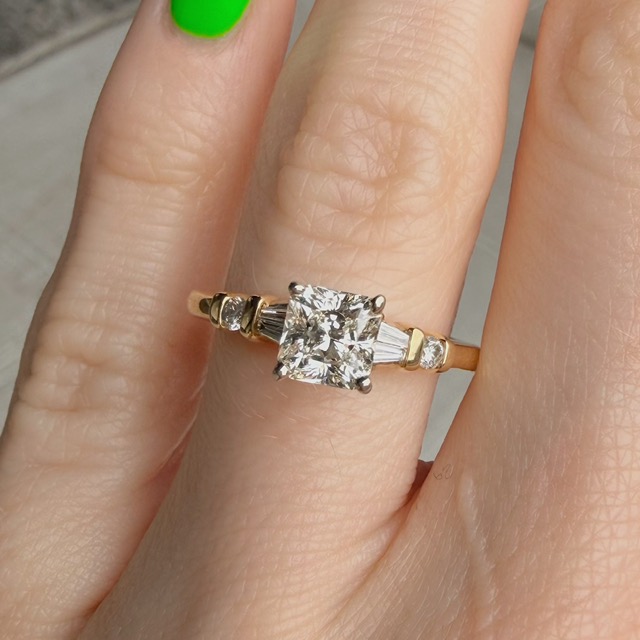 Gold engagement ring with a square-cut diamond center and baguette diamonds on the band, worn on a finger with bright green nail polish visible nearby.