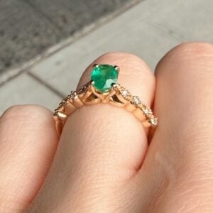 Close-up of a rose-gold ring with a green emerald centerpiece and diamond-accented band on a finger.