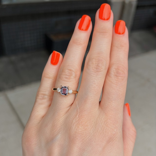 Close-up of a hand with bright orange nails wearing a gold ring featuring a purple square-cut gemstone flanked by small diamonds on each side.