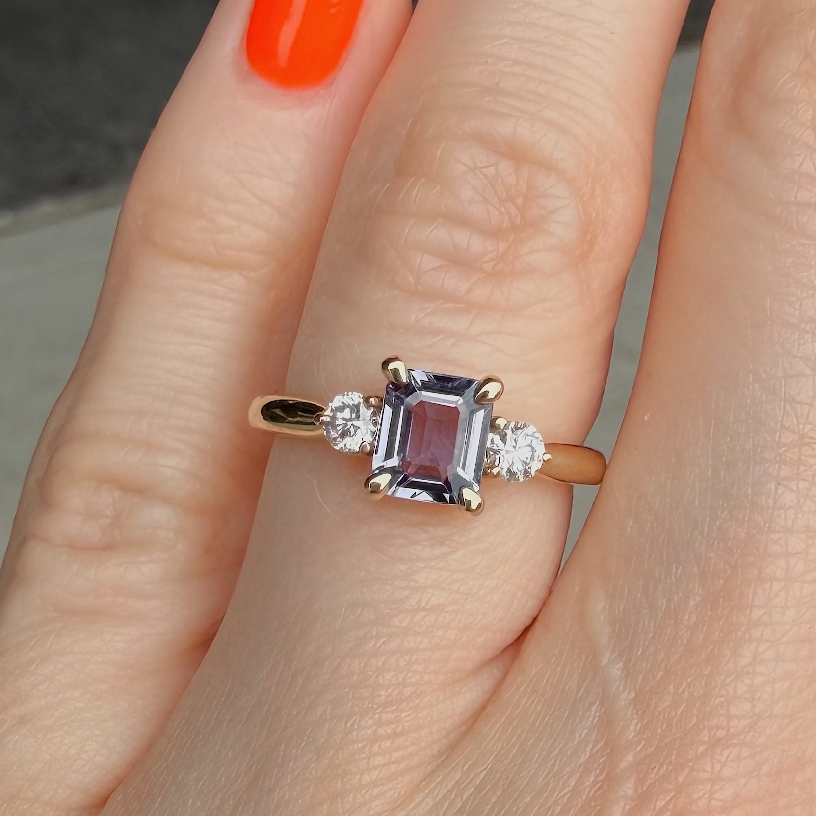 Close-up of a ring on a finger with a rectangular purple center stone flanked by two diamonds, gold setting.