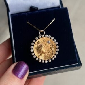 Gold coin pendant surrounded by a circle of small pearls with a tiny diamond accent, on a delicate gold chain in a navy box.
