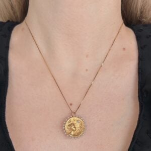 Gold circular pendant necklace with a relief design, surrounded by small bead border, resting on a woman's chest. (informative)