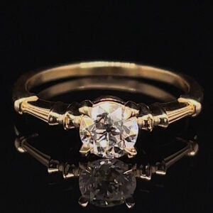 Gold solitaire engagement ring with a round diamond on a narrow band, shown on a black reflective surface.