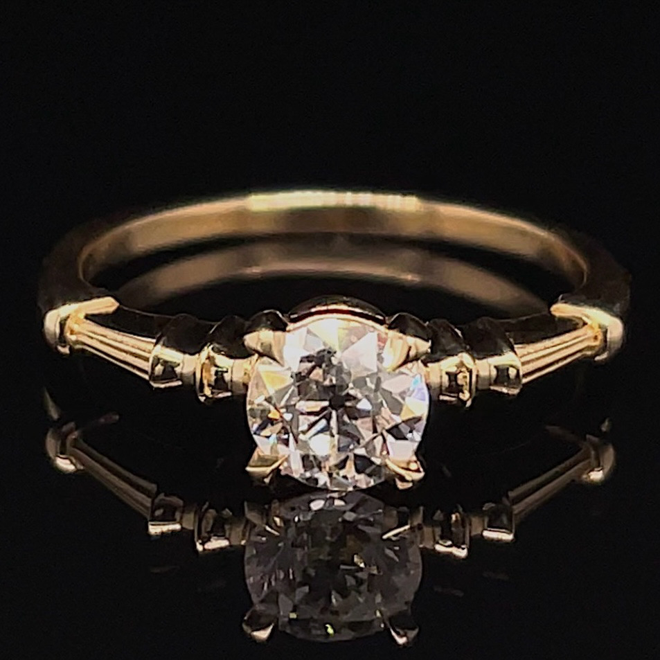 Gold solitaire engagement ring with a round diamond on a narrow band, shown on a black reflective surface.