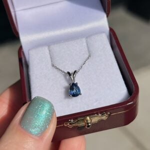 Blue teardrop pendant necklace in a silver setting on a delicate chain, placed in an open burgundy jewelry box with white velvet lining on a person's hand.