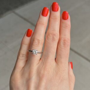 Close-up of a hand with bright red nail polish wearing a solitaire diamond engagement ring on the ring finger.
