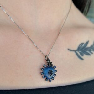 Silver necklace with a blue heart pendant surrounded by small blue stones, resting on the wearer's chest; a leaf tattoo is visible on the right.