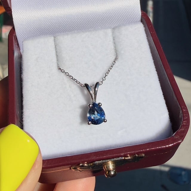 Blue teardrop pendant necklace on a silver chain inside a white padded jewelry box, held by a hand with yellow nail polish.”] ,