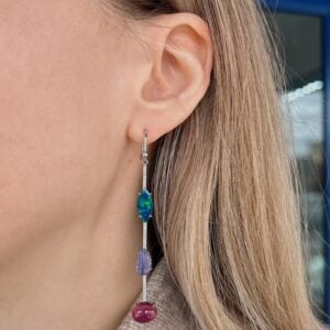 Close-up of an ear with a long silver dangle earring featuring three gemstones: blue-green oval, purple leaf, and pink-red bottom bead.