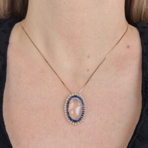 Gold chain necklace with an oval pink gemstone surrounded by small pearls and blue accents on the chest area.