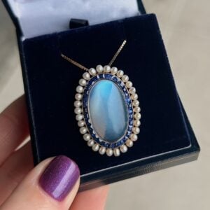 Oval blue cabochon pendant necklace in a pearl and blue-stone surround, displayed in a dark jewelry box with a gold chain.",