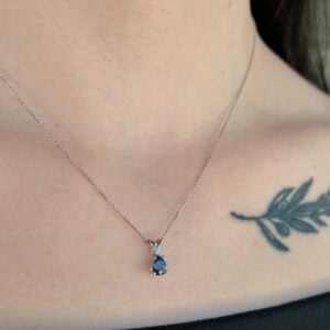 Close-up of a silver chain necklace with a blue teardrop pendant resting on the collarbone, with a leaf tattoo visible on the right shoulder.