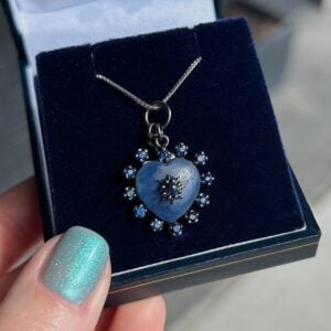 Heart-shaped blue pendant with small blue stones around it, on a silver chain in a dark blue jewelry box.