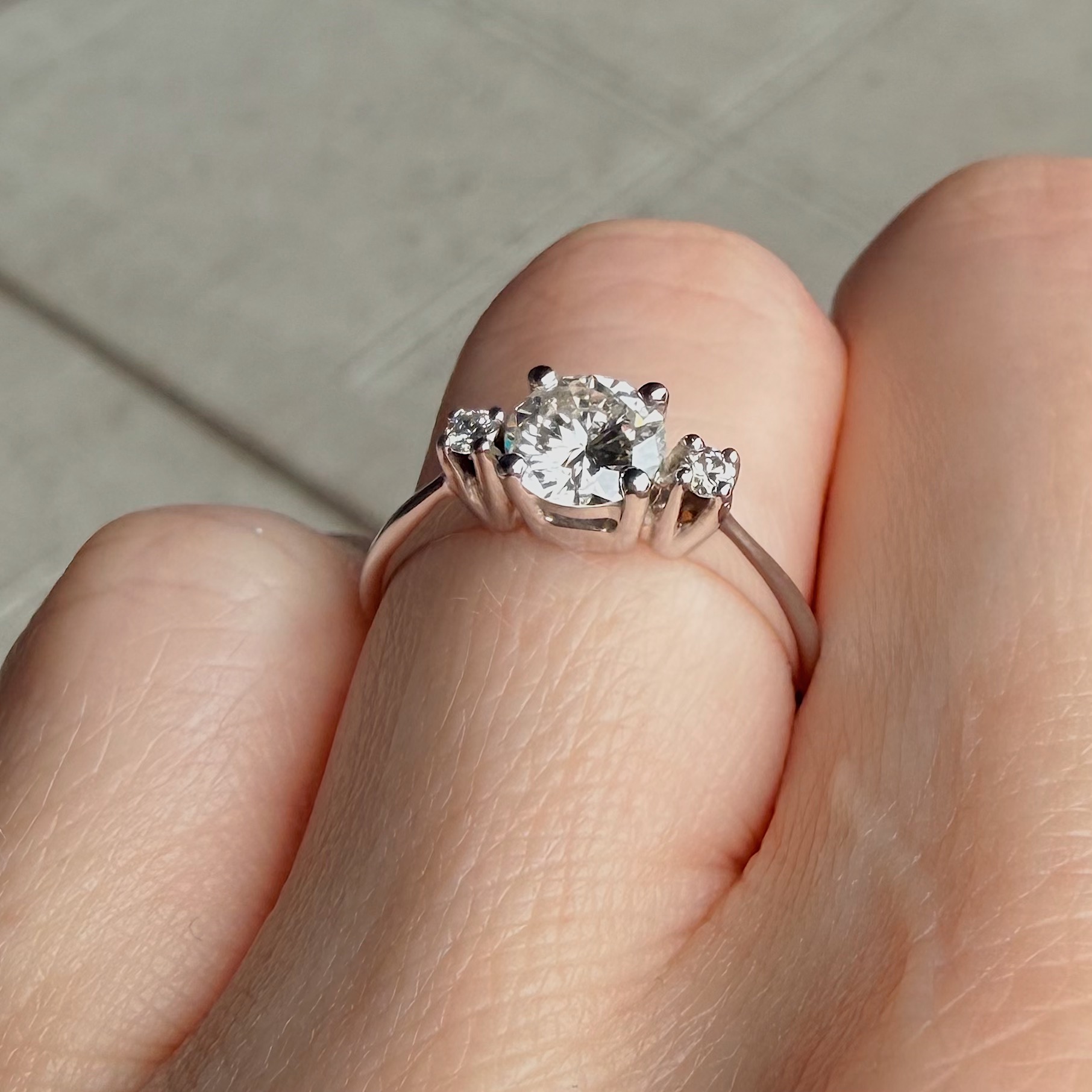 Close-up of a diamond engagement ring being held between fingers, featuring a large central stone flanked by two smaller stones.