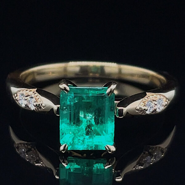 Emerald-cut green emerald ring with diamond-accented band on a polished metal band, against a black reflective surface.