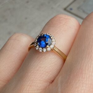 Gold ring with oval blue sapphire surrounded by a halo of small diamonds on a finger.