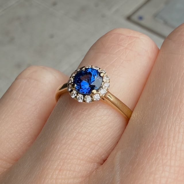 Gold ring with oval blue sapphire surrounded by a halo of small diamonds on a finger.