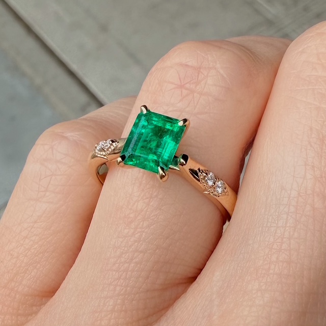 Gold ring with a large emerald-cut green emerald flanked by two small diamond-studded bands on a finger.