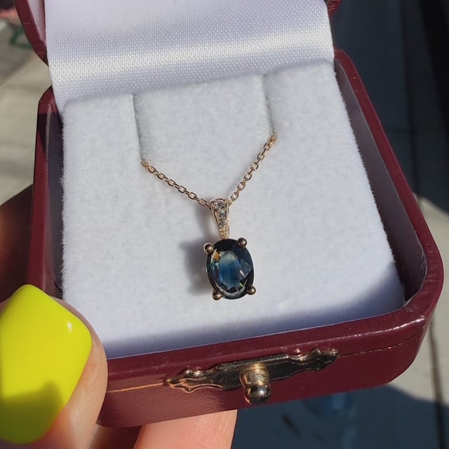 Gold chain necklace with oval blue gemstone pendant in a burgundy jewelry box lined with white foam, photographed from above.